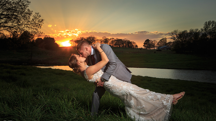 The bride and groom kissing in a field with the sun setting in the background.