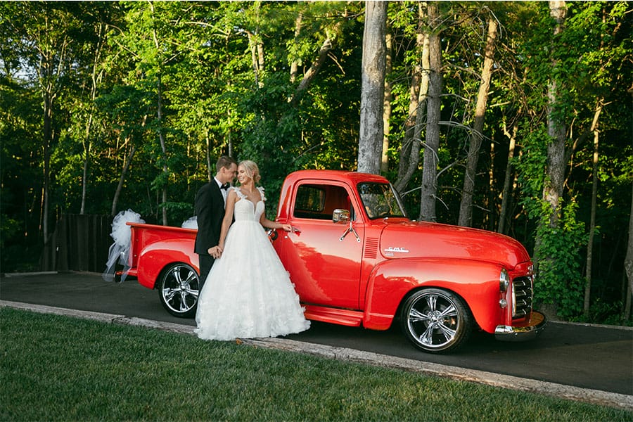 The bride and groom standing beside a red truck.